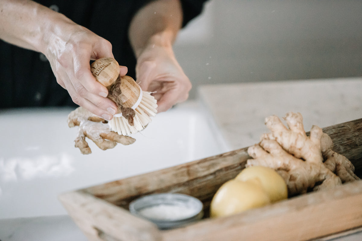 Person scrubbing ginger root with a Clean Home wooden bamboo dish brush over a sink, with a wooden tray containing ginger and lemons in the foreground. Bamboo Dish Brush from A Clean Home Company