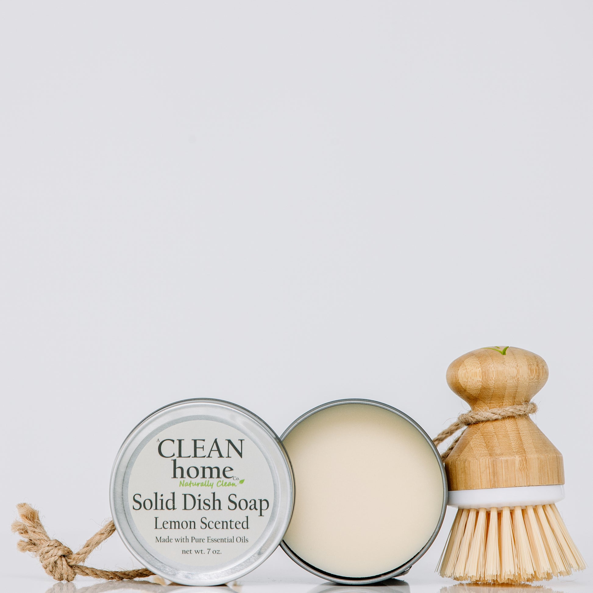 A Clean Home Company Solid Dish Soap Lemon Scented - An open tin container of solid dish soap next to a wooden dish brush with a rope loop, placed on a white surface."